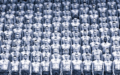 A large group of uniformed individuals arranged in a tightly packed, organized formation. Each person is wearing identical attire, consisting of a buttoned shirt and glasses. The monochromatic color scheme emphasizes the uniformity and regimentation of the scene.