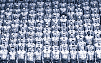 A large group of uniformed individuals arranged in a tightly packed, organized formation. Each person is wearing identical attire, consisting of a buttoned shirt and glasses. The monochromatic color scheme emphasizes the uniformity and regimentation of the scene.