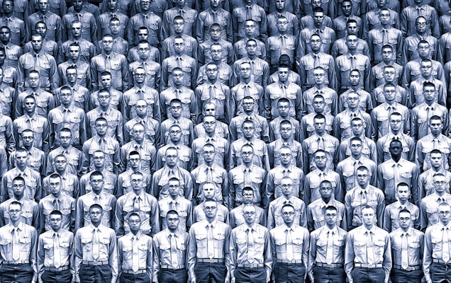 A large group of uniformed individuals arranged in a tightly packed, organized formation. Each person is wearing identical attire, consisting of a buttoned shirt and glasses. The monochromatic color scheme emphasizes the uniformity and regimentation of the scene.