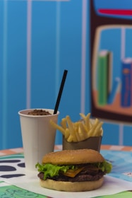 A burger with lettuce, cheese, and patty served on a sesame seed bun is placed on a table. Next to it, a portion of crispy fries is in a brown container, accompanied by a cold beverage with a straw in a white cup. The background features a blurred blue-striped wall with a bookshelf.
