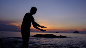 A silhouette of a person in a pose resembling Tai Chi or a meditative gesture stands against a serene backdrop of the ocean during sunset. The sky is awash with gradient hues of orange, purple, and blue, creating a calming ambiance. The silhouette is positioned near some rocks in the water, adding depth to the scene.