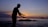 A silhouette of a person in a pose resembling Tai Chi or a meditative gesture stands against a serene backdrop of the ocean during sunset. The sky is awash with gradient hues of orange, purple, and blue, creating a calming ambiance. The silhouette is positioned near some rocks in the water, adding depth to the scene.