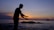 A silhouette of a person in a pose resembling Tai Chi or a meditative gesture stands against a serene backdrop of the ocean during sunset. The sky is awash with gradient hues of orange, purple, and blue, creating a calming ambiance. The silhouette is positioned near some rocks in the water, adding depth to the scene.