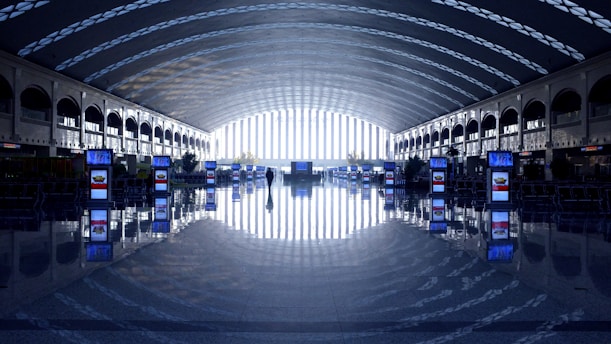 A spacious, modern airport terminal features a long, curved ceiling with a pattern of light and shadow, providing an airy and futuristic ambiance. Rows of digital displays are placed symmetrically along the reflective polished floor, while a single person walks toward a bright exit at the far end.