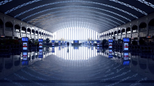 A spacious, modern airport terminal features a long, curved ceiling with a pattern of light and shadow, providing an airy and futuristic ambiance. Rows of digital displays are placed symmetrically along the reflective polished floor, while a single person walks toward a bright exit at the far end.