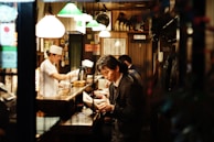 The image depicts the interior of a small, cozy restaurant with a warm ambiance. A man in a suit is sitting at the counter, reading a menu or book, while a chef, dressed in a white uniform and hat, is busy preparing food behind the counter. The restaurant is dimly lit, mainly illuminated by several hanging lamps with green and white shades. The walls are decorated with various posters and signs, adding to the intimate setting.