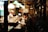 The image depicts the interior of a small, cozy restaurant with a warm ambiance. A man in a suit is sitting at the counter, reading a menu or book, while a chef, dressed in a white uniform and hat, is busy preparing food behind the counter. The restaurant is dimly lit, mainly illuminated by several hanging lamps with green and white shades. The walls are decorated with various posters and signs, adding to the intimate setting.