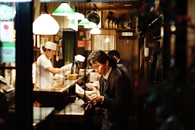 The image depicts the interior of a small, cozy restaurant with a warm ambiance. A man in a suit is sitting at the counter, reading a menu or book, while a chef, dressed in a white uniform and hat, is busy preparing food behind the counter. The restaurant is dimly lit, mainly illuminated by several hanging lamps with green and white shades. The walls are decorated with various posters and signs, adding to the intimate setting.