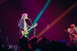A musician with long hair plays a bass guitar while singing into a microphone on stage. The stage is illuminated with colorful lights in shades of pink, blue, and orange, creating a vibrant atmosphere. Another musician is visible in the background, also performing.