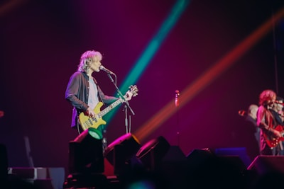 A musician with long hair plays a bass guitar while singing into a microphone on stage. The stage is illuminated with colorful lights in shades of pink, blue, and orange, creating a vibrant atmosphere. Another musician is visible in the background, also performing.