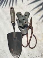A collection of gardening tools is arranged on a textured surface. The tools include a metal trowel with a wooden handle, a pair of rusted metal pruners, and several grey metallic brackets. Shadows of leaves are cast across the surface, creating a pattern.