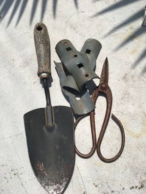 A collection of gardening tools is arranged on a textured surface. The tools include a metal trowel with a wooden handle, a pair of rusted metal pruners, and several grey metallic brackets. Shadows of leaves are cast across the surface, creating a pattern.