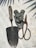 A collection of gardening tools is arranged on a textured surface. The tools include a metal trowel with a wooden handle, a pair of rusted metal pruners, and several grey metallic brackets. Shadows of leaves are cast across the surface, creating a pattern.