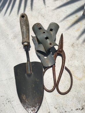 A collection of gardening tools is arranged on a textured surface. The tools include a metal trowel with a wooden handle, a pair of rusted metal pruners, and several grey metallic brackets. Shadows of leaves are cast across the surface, creating a pattern.