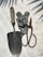 A collection of gardening tools is arranged on a textured surface. The tools include a metal trowel with a wooden handle, a pair of rusted metal pruners, and several grey metallic brackets. Shadows of leaves are cast across the surface, creating a pattern.