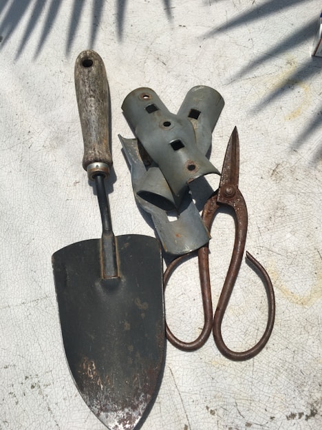 A collection of gardening tools is arranged on a textured surface. The tools include a metal trowel with a wooden handle, a pair of rusted metal pruners, and several grey metallic brackets. Shadows of leaves are cast across the surface, creating a pattern.