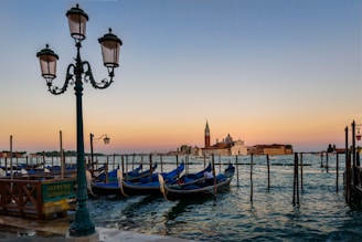 A scenic view of a waterfront with gondolas moored to wooden posts. In the background, there is an island with a prominent church and bell tower. A vintage-style lamppost and a sign that reads 'Servizio Gondole' are on the left side. The sky is a gradient of colors, transitioning from warm orange near the horizon to cooler blue hues as it ascends.