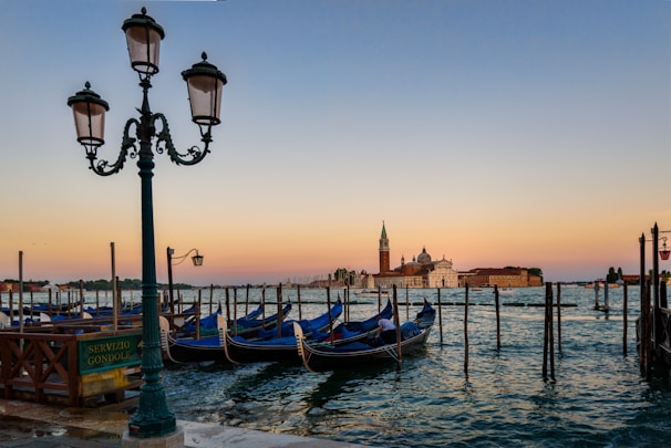 A scenic view of a waterfront with gondolas moored to wooden posts. In the background, there is an island with a prominent church and bell tower. A vintage-style lamppost and a sign that reads 'Servizio Gondole' are on the left side. The sky is a gradient of colors, transitioning from warm orange near the horizon to cooler blue hues as it ascends.