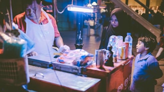 A street vendor in a red shirt and white apron is preparing food at a stall, illuminated by a bright light. A woman in a black hijab and a child in a blue shirt are standing on the other side of the stall, looking at the vendor. Various items such as a water bottle, a can, and cups are arranged on the countertop.