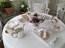 An elegantly set table with a white tablecloth adorned with floral-patterned china, a silver teapot, silver bowls, and a cake stand filled with various pastries. A glass cloche with a pretzel or pastry beneath it is also present. The table is set for four, with tea cups and saucers placed at each setting. The setting includes a white lace-covered armchair with a floral cushion, and potted plants and additional furniture in the background.