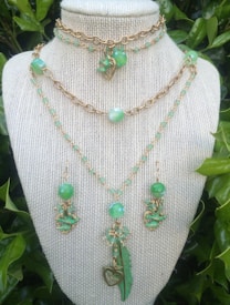 A set of intricately designed jewelry featuring green beads and gold chains is displayed on a mannequin. The set includes two necklaces and a pair of earrings. The necklaces are adorned with green beads, gold chain links, and pendants shaped like hearts and feathers. The earrings have similar green bead and gold accents.