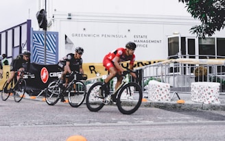 Several cyclists wearing helmets are racing in an outdoor event. The riders are on a paved path, and the area is bordered by barriers and banners advertising different brands. Some people are observing the race from behind the barricades. The background includes a building with the words 'Greenwich Peninsula Estate Management Office' displayed.