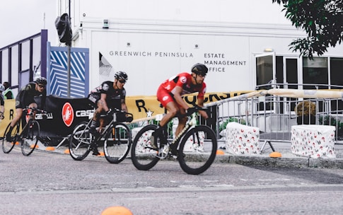 Several cyclists wearing helmets are racing in an outdoor event. The riders are on a paved path, and the area is bordered by barriers and banners advertising different brands. Some people are observing the race from behind the barricades. The background includes a building with the words 'Greenwich Peninsula Estate Management Office' displayed.