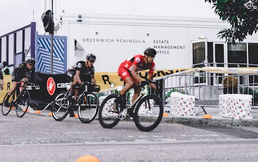 Several cyclists wearing helmets are racing in an outdoor event. The riders are on a paved path, and the area is bordered by barriers and banners advertising different brands. Some people are observing the race from behind the barricades. The background includes a building with the words 'Greenwich Peninsula Estate Management Office' displayed.