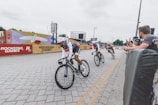 Cyclists are engaged in a racing event along a paved street, with spectators watching from behind barriers. The area is decorated with vibrant artwork, and branded banners are visible.