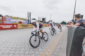 Cyclists are engaged in a racing event along a paved street, with spectators watching from behind barriers. The area is decorated with vibrant artwork, and branded banners are visible.
