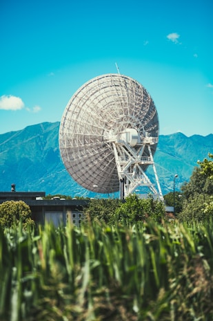 A large satellite dish stands prominently in a natural setting with lush greenery in the foreground. Behind the dish, a range of mountains is visible under a clear blue sky, suggesting a remote and serene environment.