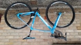 A blue bicycle is mounted upside down against a brick wall. The bike features a lightweight frame and thin wheels, suitable for road cycling. The handlebars are wrapped in black tape, and the brand name is prominently displayed on the frame.