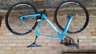 A blue bicycle is mounted upside down against a brick wall. The bike features a lightweight frame and thin wheels, suitable for road cycling. The handlebars are wrapped in black tape, and the brand name is prominently displayed on the frame.