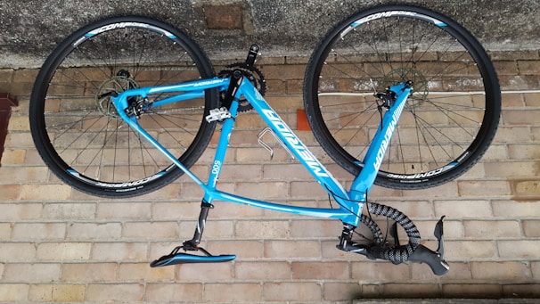A blue bicycle is mounted upside down against a brick wall. The bike features a lightweight frame and thin wheels, suitable for road cycling. The handlebars are wrapped in black tape, and the brand name is prominently displayed on the frame.