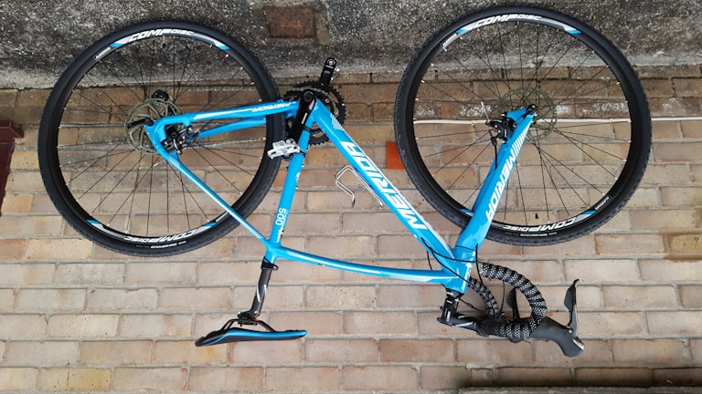 A blue bicycle is mounted upside down against a brick wall. The bike features a lightweight frame and thin wheels, suitable for road cycling. The handlebars are wrapped in black tape, and the brand name is prominently displayed on the frame.