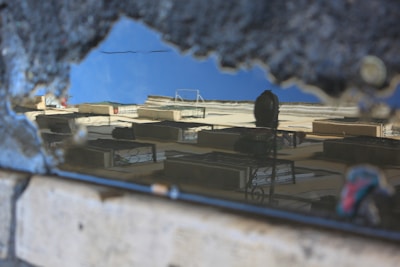 Reflection of a building in a rain puddle on a rough, textured ground surface. The building's windows and balconies are visible, with part of the clear blue sky showing in the water.