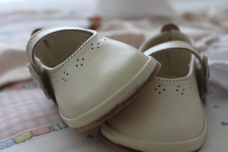 A pair of small, beige-colored, leather baby shoes with delicate perforated designs and a strap across the top. They are placed on a soft, patterned surface which appears to be fabric, possibly a blanket or clothing.
