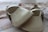 A pair of small, beige-colored, leather baby shoes with delicate perforated designs and a strap across the top. They are placed on a soft, patterned surface which appears to be fabric, possibly a blanket or clothing.