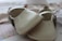 A pair of small, beige-colored, leather baby shoes with delicate perforated designs and a strap across the top. They are placed on a soft, patterned surface which appears to be fabric, possibly a blanket or clothing.