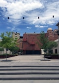 An open urban plaza features a series of concrete steps leading up to a flat paved area with trees and planters on either side. In the background, a red brick building with a dome and multiple windows stands out against a blue sky with scattered clouds. Overhead, a string of black rectangular lights stretches across the scene.