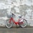 A red rental bicycle is parked against a weathered, textured brick wall with peeling white paint. The ground is an asphalt surface with patches of grass growing through cracks.