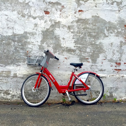 A red rental bicycle is parked against a weathered, textured brick wall with peeling white paint. The ground is an asphalt surface with patches of grass growing through cracks.