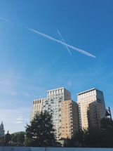 A cluster of modern, high-rise buildings under a clear blue sky with visible contrails forming an X shape. The buildings are surrounded by trees and a distant dome structure can be seen to the left. The scene is urban, with a lamppost and satellite dishes in the foreground.
