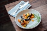 A beautifully presented gourmet dish with colorful roasted vegetables, including carrots and orange segments, garnished with herbs and seeds, served on a white plate. The plate is set on a wooden table with a white cloth napkin and silver cutlery beside it.