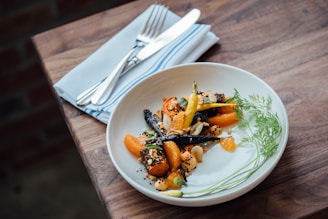 A beautifully presented gourmet dish with colorful roasted vegetables, including carrots and orange segments, garnished with herbs and seeds, served on a white plate. The plate is set on a wooden table with a white cloth napkin and silver cutlery beside it.