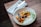 A beautifully presented gourmet dish with colorful roasted vegetables, including carrots and orange segments, garnished with herbs and seeds, served on a white plate. The plate is set on a wooden table with a white cloth napkin and silver cutlery beside it.