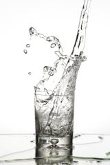 Water splashes dynamically as it is poured into a clear glass, with droplets suspended in mid-air and a rippling surface below.
