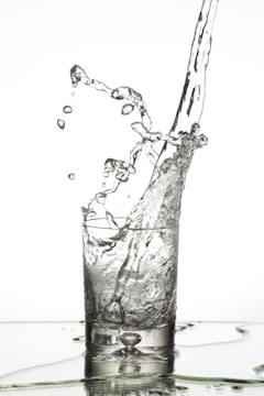 Water splashes dynamically as it is poured into a clear glass, with droplets suspended in mid-air and a rippling surface below.