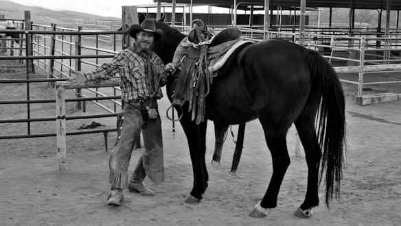 A person dressed in cowboy attire, including a hat and chaps, stands next to a saddled horse in a ranch setting. The background displays a series of fenced paddocks and stables, suggesting an equestrian or livestock environment. The overall scene gives an impression of rural or western life.