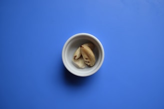 A small white bowl contains Brazil nuts, set against a solid blue background. The nuts are light brown with dark brown markings.